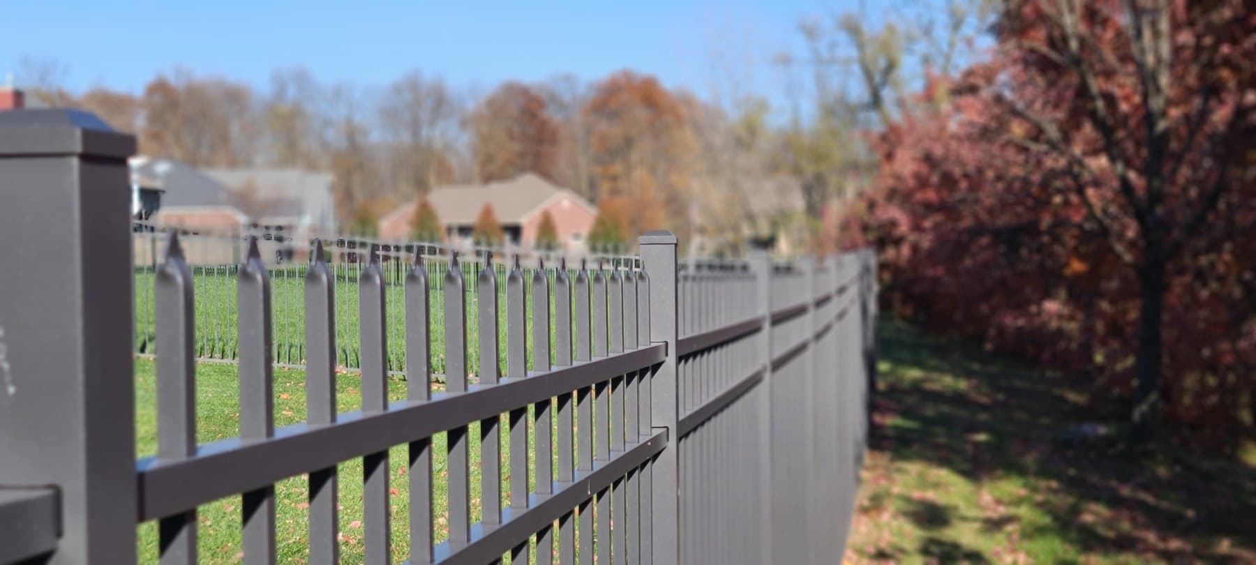 An aluminum fence follows a curving boundary beside autumn trees.