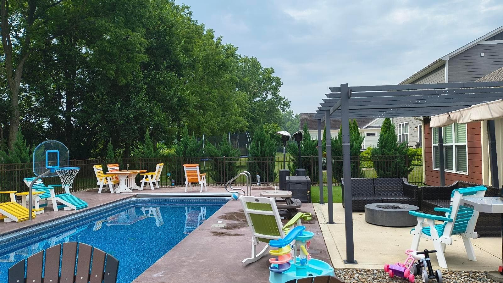 A black fence borders a pool with colorful chairs and a shaded seating area.