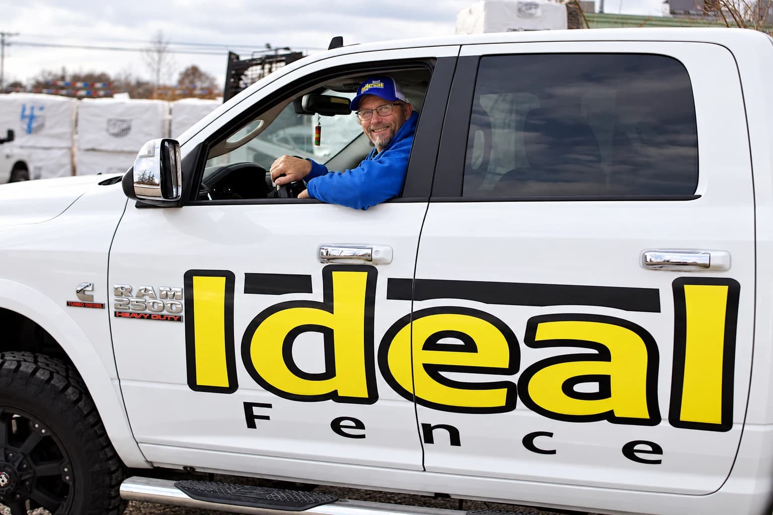 Ideal Fence team member in a blue company sweatshirt at the wheel of a white work truck with a large Ideal Fence door logo.