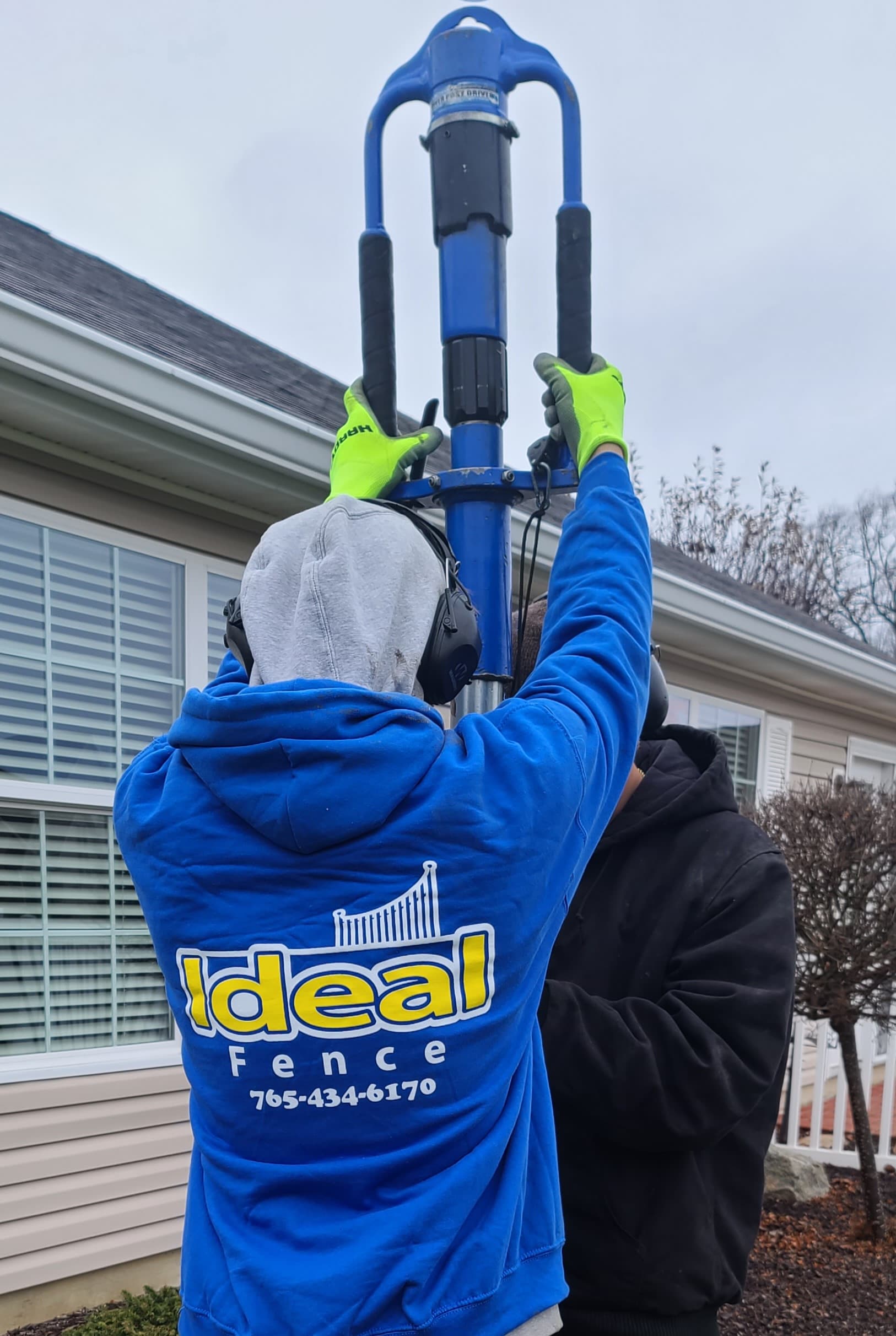 Ideal Fence installer operating a powered post driver over a fence post beside a house.