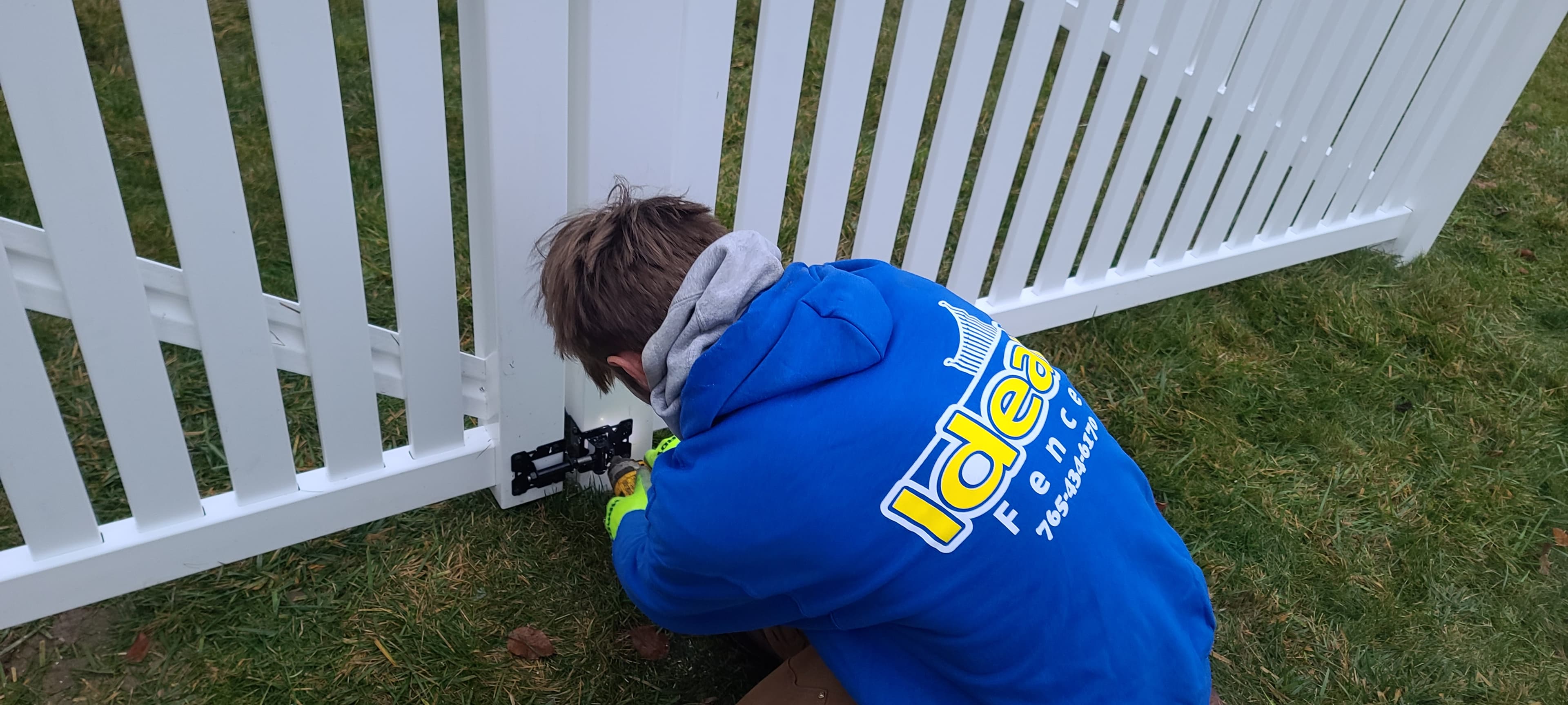 Ideal Fence installer adjusting black hardware on a white vinyl picket gate with open vertical pickets.