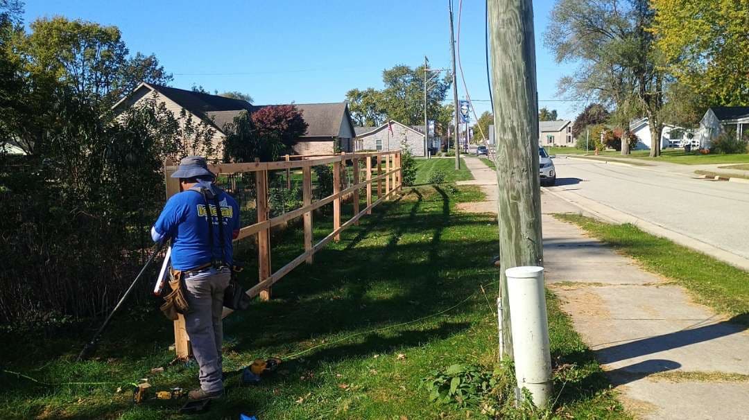 Ideal Fence installer working beside a wood fence frame with exposed posts and horizontal rails along a sidewalk.