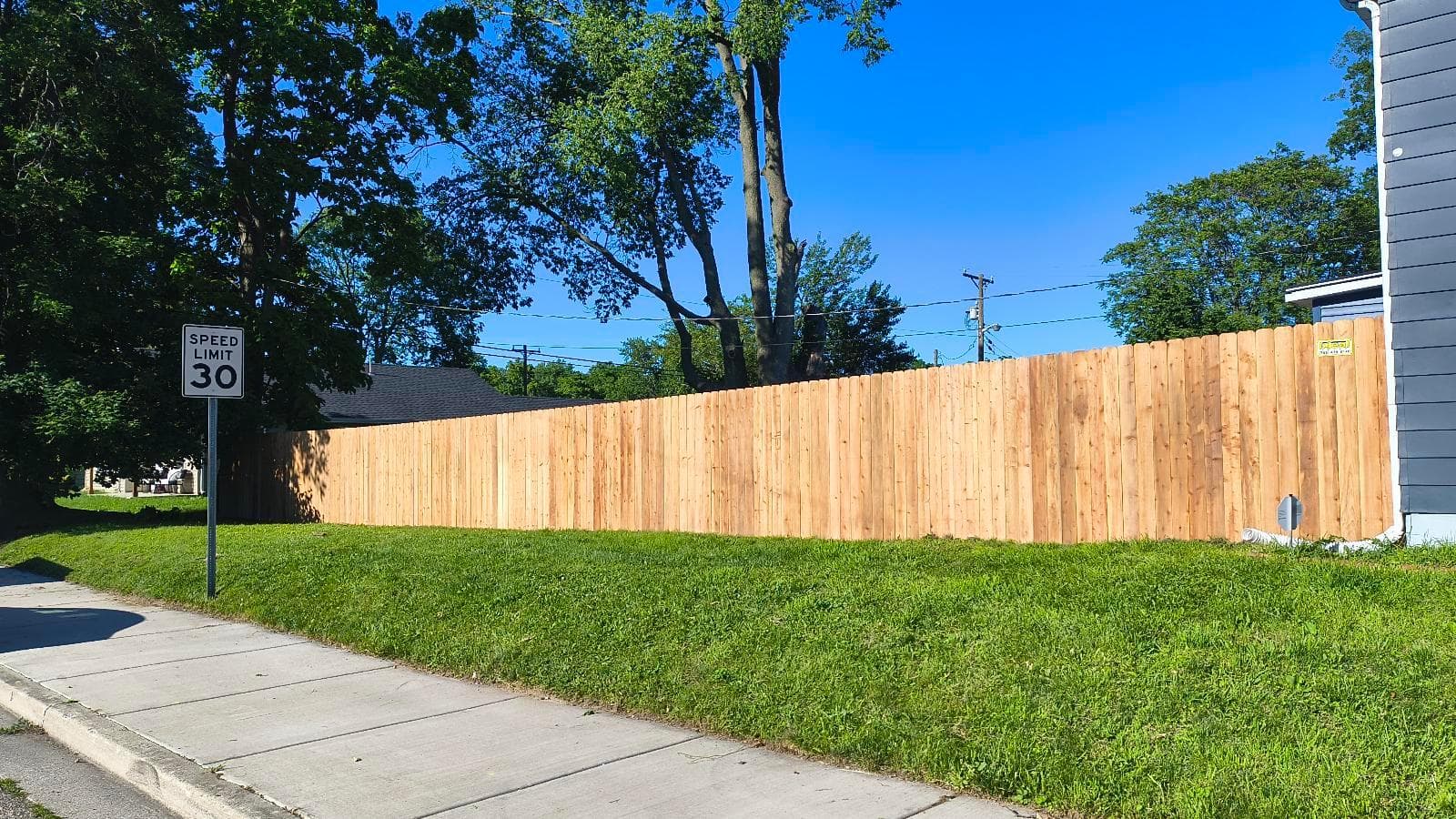 Vertical wood dog-ear privacy fence following a grassy bank beside a sidewalk and a shaded residential yard.